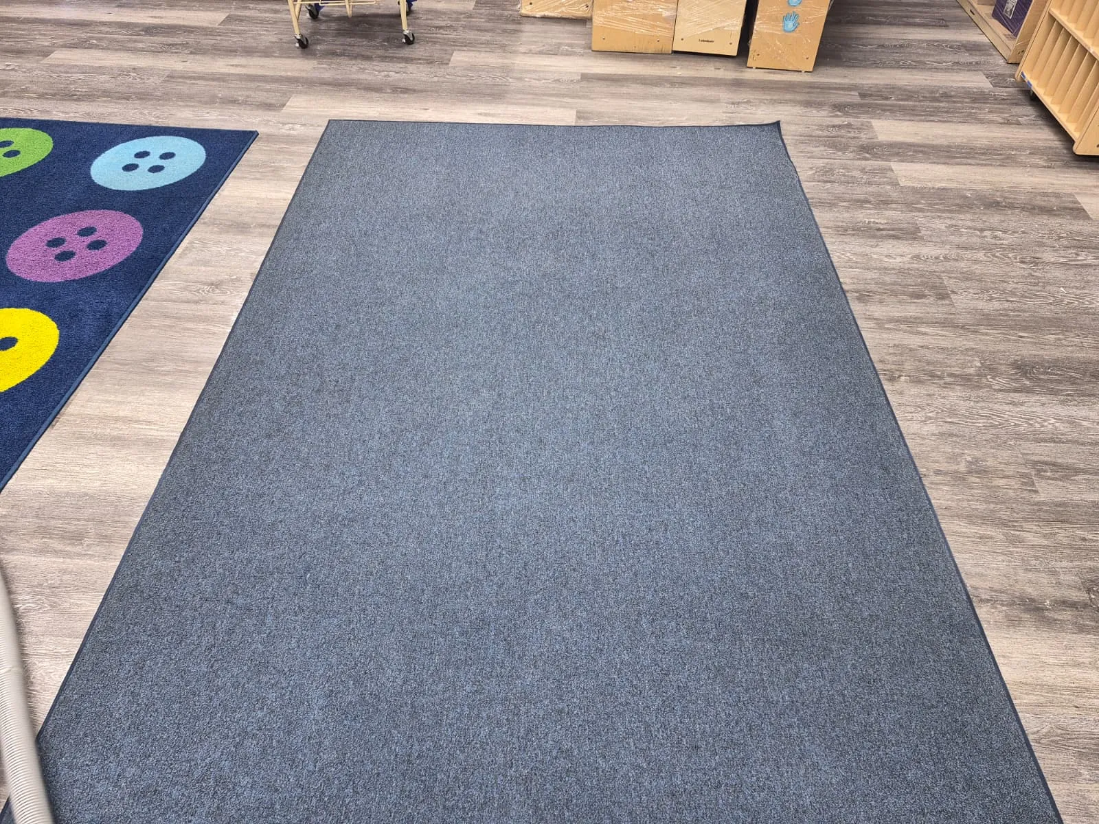 A freshly cleaned grey commercial carpet laid over a wood floor in a classroom, next to a patterned children’s rug