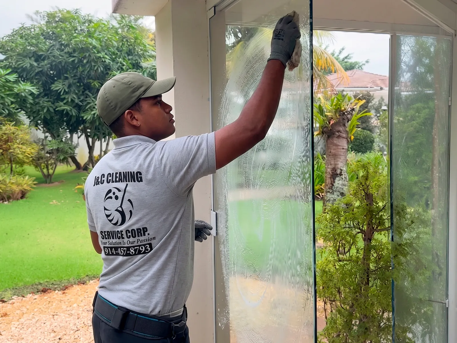 A J&C Cleaning Services technician in a branded polo and work gloves washing the glass of an entrance door